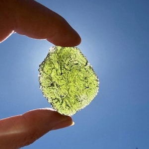 Puede incluir: Una gema verde translúcida, de forma irregular, se sostiene entre los dedos contra un cielo azul brillante. La piedra tiene una superficie texturizada y parece estar iluminada por la luz solar. La imagen resalta la belleza natural del mineral.