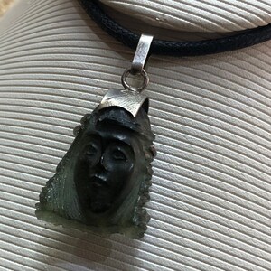 May include: A silver chain necklace with a green stone pendant featuring a carved face. The pendant is shaped like a teardrop and has a detailed design.