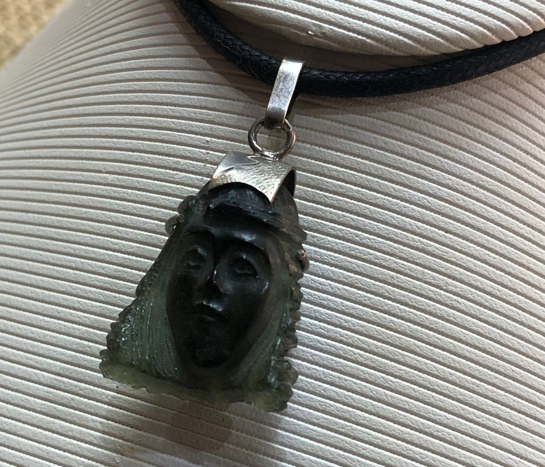 May include: A silver chain necklace with a green stone pendant featuring a carved face. The pendant is shaped like a teardrop and has a detailed design.