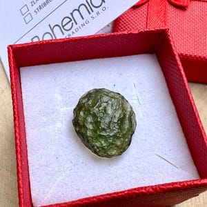 Könnte beinhalten: Ein grüner, rauer und strukturierter Edelstein in einer roten Samtbox. Der Edelstein ist ein Moldavit, eine Art Tektit.