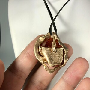 May include: A gold pendant necklace with a large amber stone set in a textured, abstract design. The necklace is on a black leather cord.