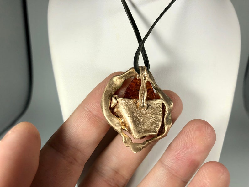 May include: A gold pendant necklace with a large amber stone set in a textured, abstract design. The necklace is on a black leather cord.