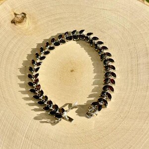 May include: A silver bracelet featuring a row of dark red gemstones set in a leaf-like design. The bracelet has a clasp fastening.