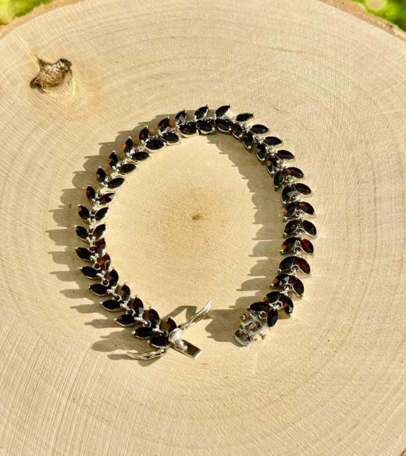 May include: A silver bracelet featuring a row of dark red gemstones set in a leaf-like design. The bracelet has a clasp fastening.
