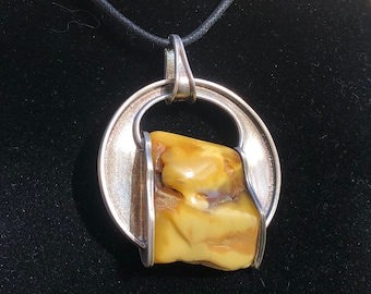 May include: A silver pendant necklace with a large, irregular amber stone set in a circular frame. The amber is a warm, honey color with some darker brown streaks.