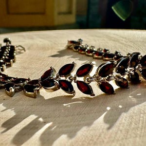 May include: A silver bracelet featuring a row of dark red marquise-shaped gemstones. The bracelet is crafted from a delicate chain and has a clasp fastening.