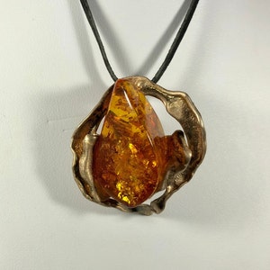May include: A bronze pendant with a large amber stone set in a swirling, abstract design. The pendant is attached to a black leather cord.