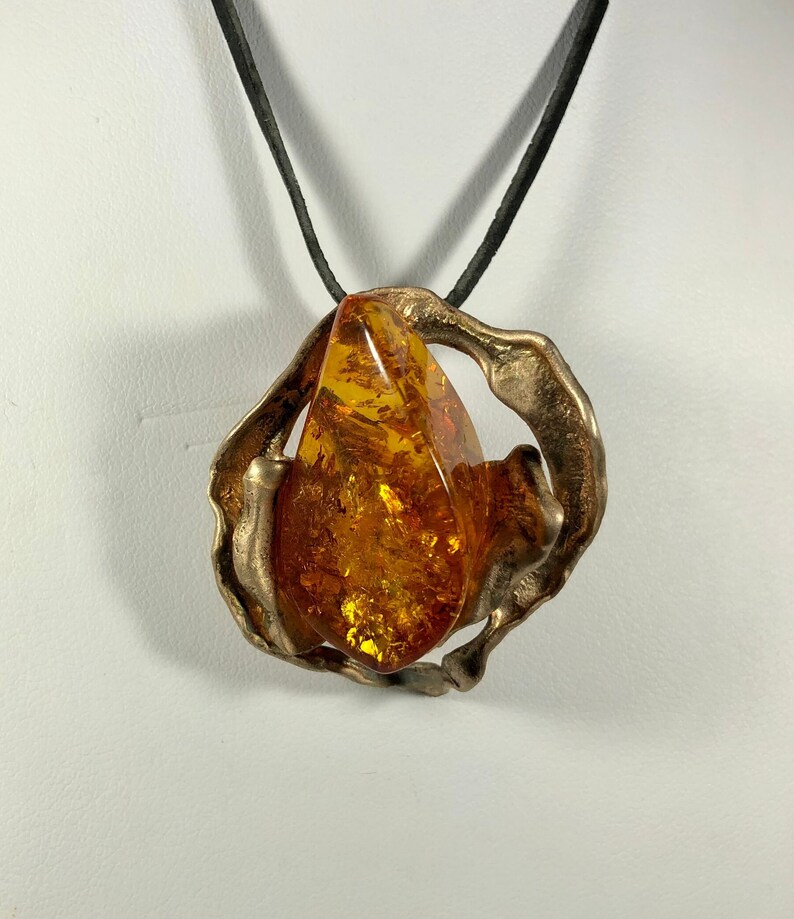 May include: A bronze pendant with a large amber stone set in a swirling, abstract design. The pendant is attached to a black leather cord.