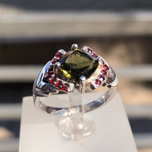 May include: A silver ring with a green gemstone surrounded by red gemstones.