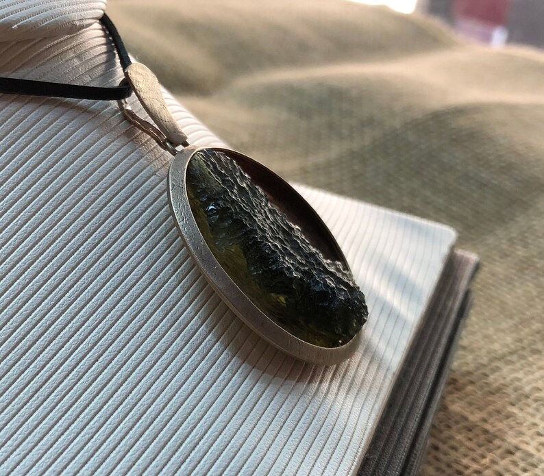 May include: A silver pendant with a round green and black stone. The stone has a rough, textured surface.