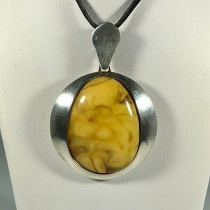 May include: A silver pendant with a large, oval-shaped amber stone. The amber is a warm, honey color with swirls and inclusions. The pendant is on a black leather cord.
