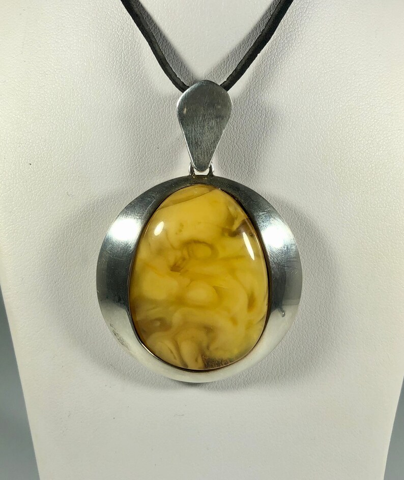 May include: A silver pendant with a large, oval-shaped amber stone. The amber is a warm, honey color with swirls and inclusions. The pendant is on a black leather cord.