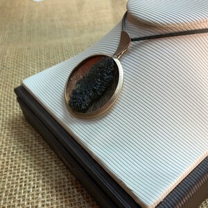 May include: A round pendant necklace with a dark green and brown stone set in a silver-toned metal frame. The necklace is on a black cord and is displayed on a white stand.