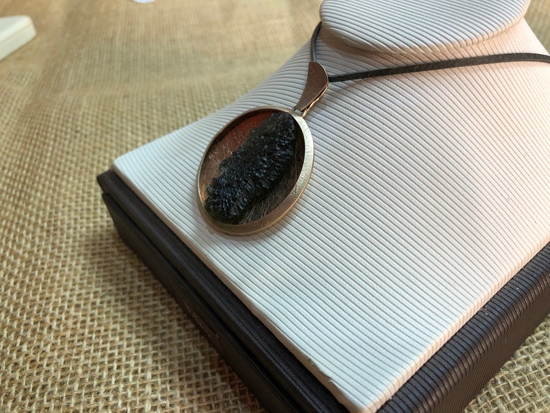 May include: A round pendant necklace with a dark green and brown stone set in a silver-toned metal frame. The necklace is on a black cord and is displayed on a white stand.