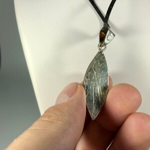 May include: Silver pendant with a textured, leaf-shaped design. The pendant is attached to a black cord.