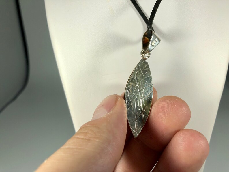 May include: Silver pendant with a textured, leaf-shaped design. The pendant is attached to a black cord.