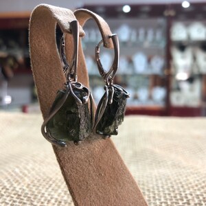 May include: A pair of silver earrings with green gemstone drops. The earrings are on a brown display stand.