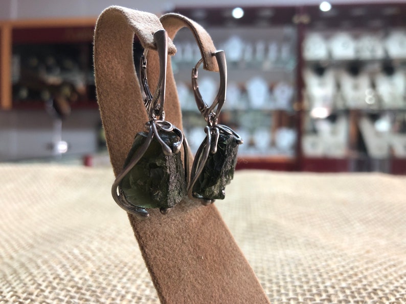 May include: A pair of silver earrings with green gemstone drops. The earrings are on a brown display stand.