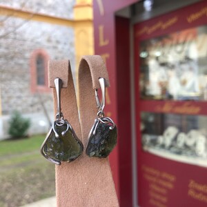 May include: A pair of silver earrings with green gemstone drops. The earrings are displayed on a brown velvet stand.