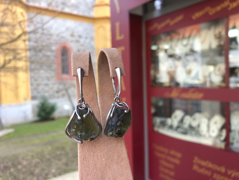 May include: A pair of silver earrings with green gemstone drops. The earrings are displayed on a brown velvet stand.