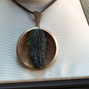 May include: A round silver pendant necklace with a black and green stone encased in resin. The stone has a rough, textured surface.