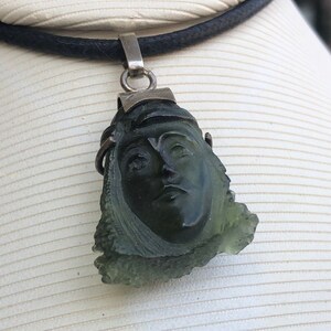 May include: A green gemstone pendant carved with a face, set in silver and hanging from a black cord.