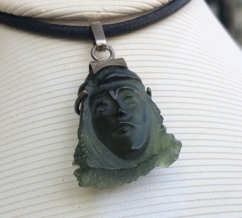 May include: A green gemstone pendant carved with a face, set in silver and hanging from a black cord.