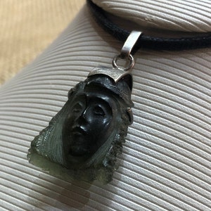 May include: A silver pendant necklace with a dark green face carved into the pendant. The necklace is on a black cord.