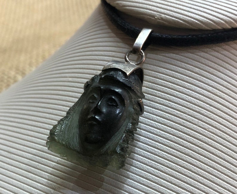 May include: A silver pendant necklace with a dark green face carved into the pendant. The necklace is on a black cord.