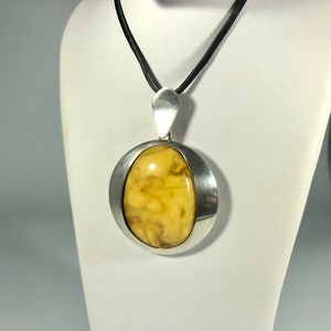 May include: A silver pendant with a round, yellow amber stone set in a silver bezel. The pendant is on a black cord.