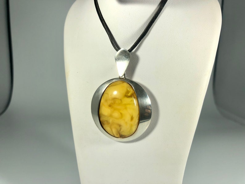 May include: A silver pendant with a round, yellow amber stone set in a silver bezel. The pendant is on a black cord.