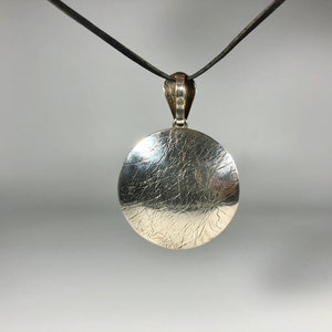 May include: A silver pendant with a textured, circular design hangs from a black cord.