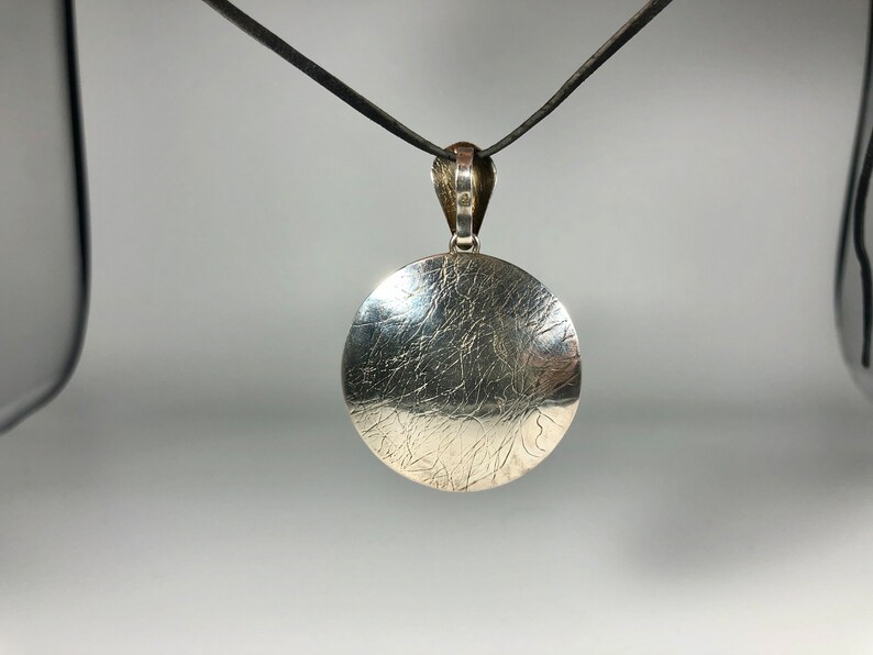 May include: A silver pendant with a textured, circular design hangs from a black cord.