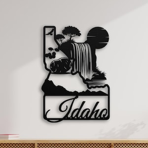 May include: Black metal wall art depicting the state of Idaho with a waterfall, trees, and a sun. The word "Idaho" is written in cursive at the bottom of the artwork. The piece is mounted on a white wall.