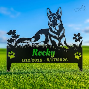 May include: A black metal garden stake featuring a silhouette of a German Shepherd dog lying in grass with flowers on each side. The name Rocky is cut out in the center above the dates 1/12/2015 - 5/17/2026, flanked by two paw prints.