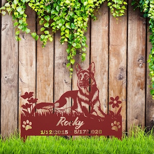 May include: A dark red metal garden stake featuring a German Shepherd dog lying in grass with flowers. The name Rocky is cut out in the center, with the dates 1/12/2015 and 5/17/2026 printed below, flanked by two paw print cutouts on the sides.