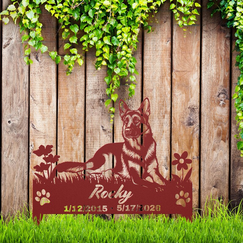 May include: A dark red metal garden stake featuring a German Shepherd dog lying in grass with flowers. The name Rocky is cut out in the center, with the dates 1/12/2015 and 5/17/2026 printed below, flanked by two paw print cutouts on the sides.
