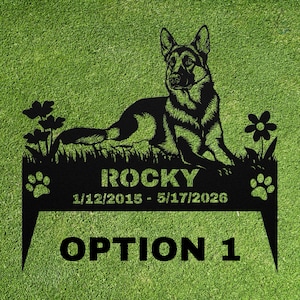 May include: A black metal garden stake featuring a German Shepherd silhouette lying in grass with flowers. The text reads ROCKY, 1/12/2015 - 5/17/2026, and OPTION 1, flanked by two paw prints. The design includes two pointed stakes at the bottom.