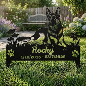 May include: A black metal garden memorial stake featuring a silhouette of a German Shepherd dog lying down, surrounded by flowers and paw prints. The text reads Rocky and the dates 1/12/2015 - 5/17/2026, centered on the rectangular base.