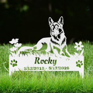 May include: A white metal garden stake featuring a cutout silhouette of a German Shepherd dog lying in grass with flowers. The base includes the name Rocky and the dates 1/12/2015 - 5/17/2026, flanked by two paw print cutouts on the left and right sides.