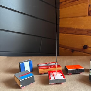 May include: A set of six small wooden boxes with different colored tops and black bottoms. The boxes are arranged in a row on a brown surface. A black incense stick is standing upright in the center of the image.