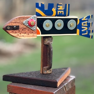 May include: A whimsical wooden fish sculpture with a brown body, black midsection, and blue and yellow accents. The sculpture features the word "LINE" and "LVAN". It is mounted on a wooden base with a handle.