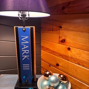 May include: A table lamp with a purple lampshade and a base featuring the text "MARK III" in white. The lamp sits on a blue surface, with a bowl of ornaments and several loose ornaments in shades of blue, green, and copper.