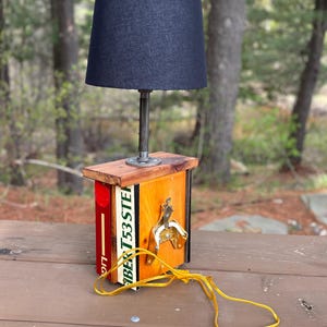 May include: A handmade lamp with a navy blue lampshade and a wooden base. The base is constructed from repurposed materials, including a beer bottle opener and a label that reads "BER T53 STEP". The lamp has a yellow power cord.