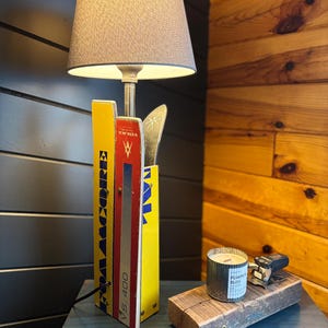 May include: A unique lamp with a gray lampshade and a base made from repurposed skis. The skis are yellow, red, and white, with the text "VOLKL" and "VS 400" visible. A candle and a small object sit on a wooden block.