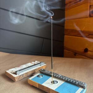 May include: A wooden incense holder with a blue and white striped design. A metal incense stick holder is attached to the top of the holder. A black incense stick is burning and releasing smoke.
