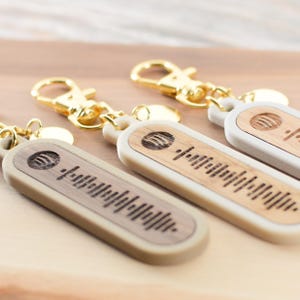 Könnte beinhalten: Drei beige Schlüsselanhänger mit goldenen Metallschließen. Jeder Schlüsselanhänger hat einen rechteckigen Holzeinsatz mit einem schwarz-weißen Barcode-Design und einen kleinen Kreis mit einem Wellendesign.