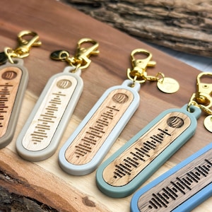 Könnte beinhalten: Fünf Holzschlüsselanhänger mit verschiedenen farbig lackierten Harzbeschichtungen. Jeder Schlüsselanhänger zeigt ein einzigartiges Schallwellendesign, das in das Holz graviert ist. Die Schlüsselanhänger sind an goldfarbenen Metallschlüsselringen befestigt.