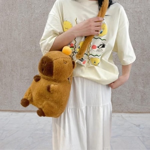 May include: A brown plush capybara-shaped crossbody bag with a long, furry strap. The bag has a round head, small ears, and tiny arms and legs. The bag is worn over a white skirt and a light yellow t-shirt with cartoon characters.