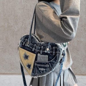 Może przedstawiać: Damska torebka w kształcie serca z dżinsu z patchworkiem. Torba posiada jasnobrązowy trójkąt z wycięciami w kształcie gwiazd i postrzępionymi krawędziami. Torba ma pasek na ramię i jest wykonana z niebieskiego dżinsu.